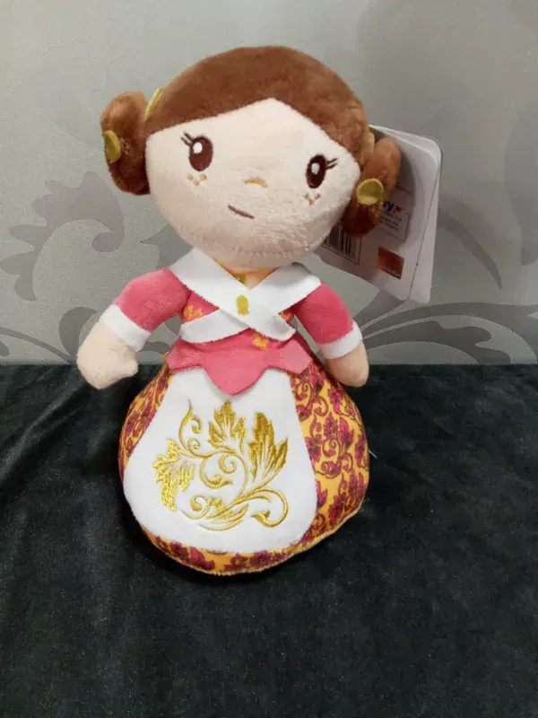 Muñeca de peluche con cabello castaño, vestido rosa y delantal blanco con bordados florales dorados.
