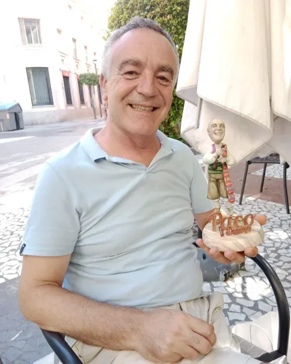 Un hombre sonriente, vestido con un polo azul claro, está sentado al aire libre y sostiene una figura de él.