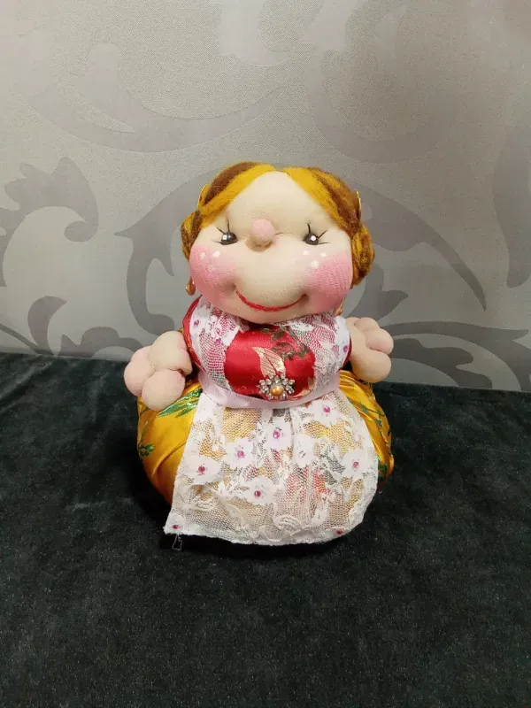 Muñeca pequeña hecha a mano con cabello rubio, mejillas rosadas y vestido floral sentada sobre un fondo gris estampado.