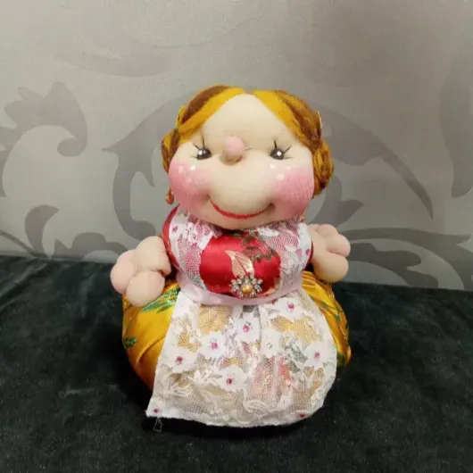 Muñeca pequeña hecha a mano con rostro sonriente, vestido rojo y blanco y cabello amarillo sobre un fondo gris estampado.