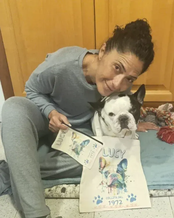 Una mujer se agacha junto a un perro pequeño que sostiene una tarjeta y un póster sobre una cama.