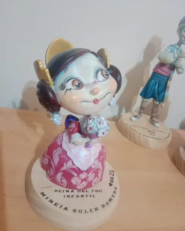 Figurita colorida de una niña con un vestido rosa sobre una repisa de madera, con otra figurita detrás de ella.