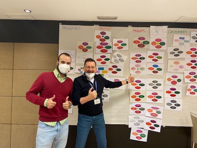 Dos hombres con máscaras hacen un gesto de aprobación con el pulgar frente a un tablero con círculos.