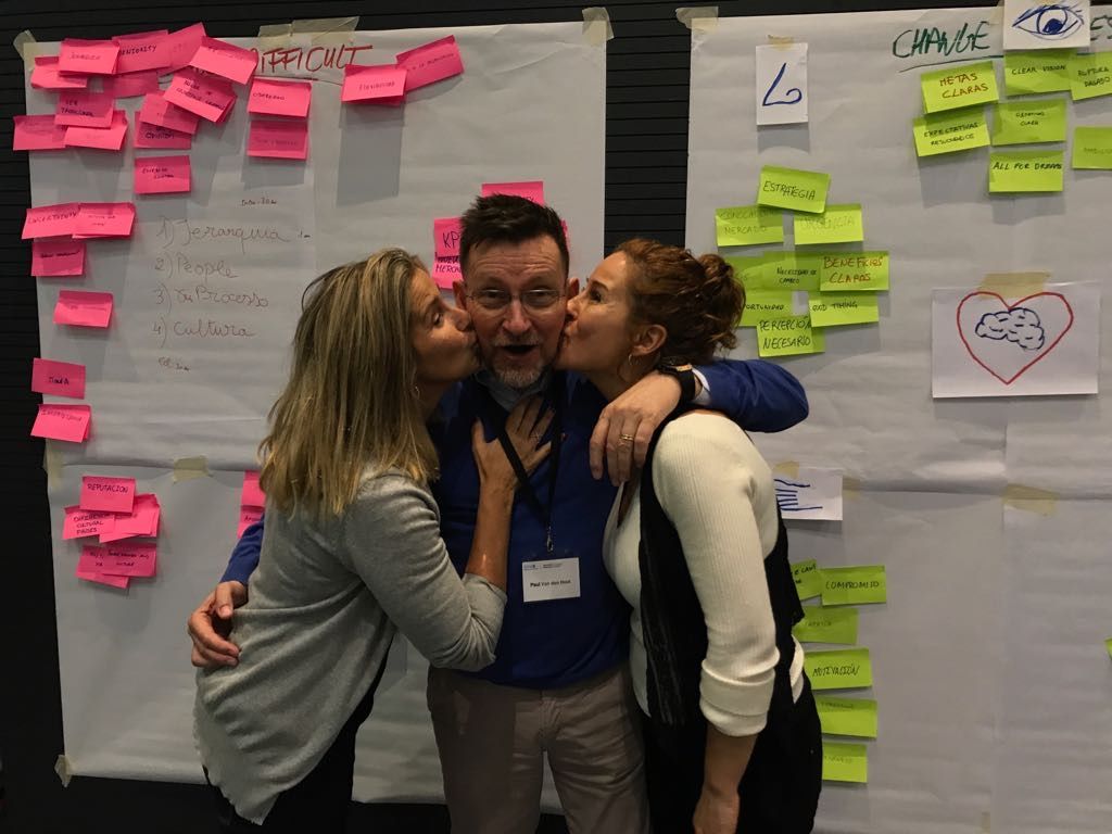 Dos mujeres besan a un hombre en la mejilla frente a un tablero con notas adhesivas.