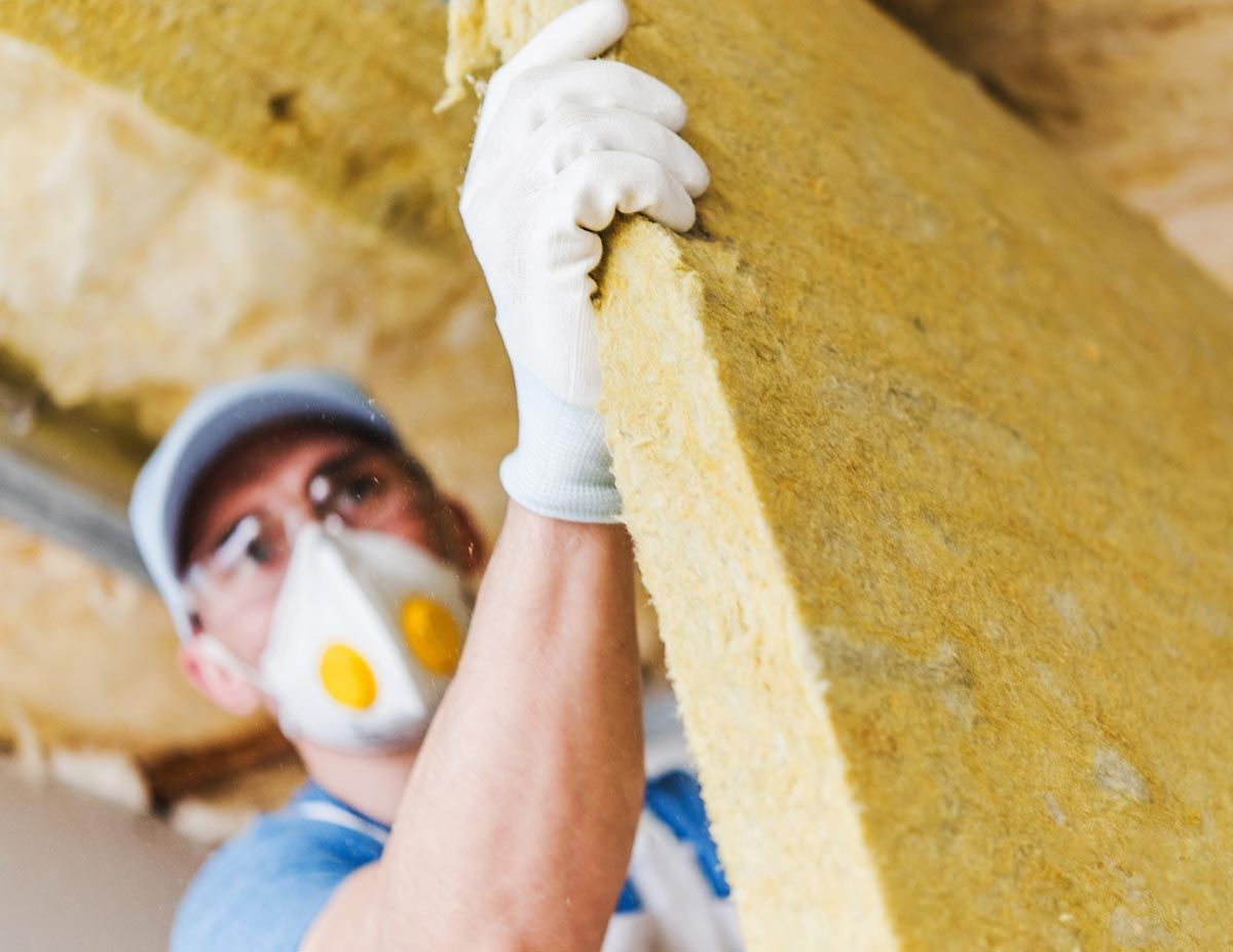 Un trabajador con mascarilla y guantes instala aislamiento amarillo en el hueco del techo