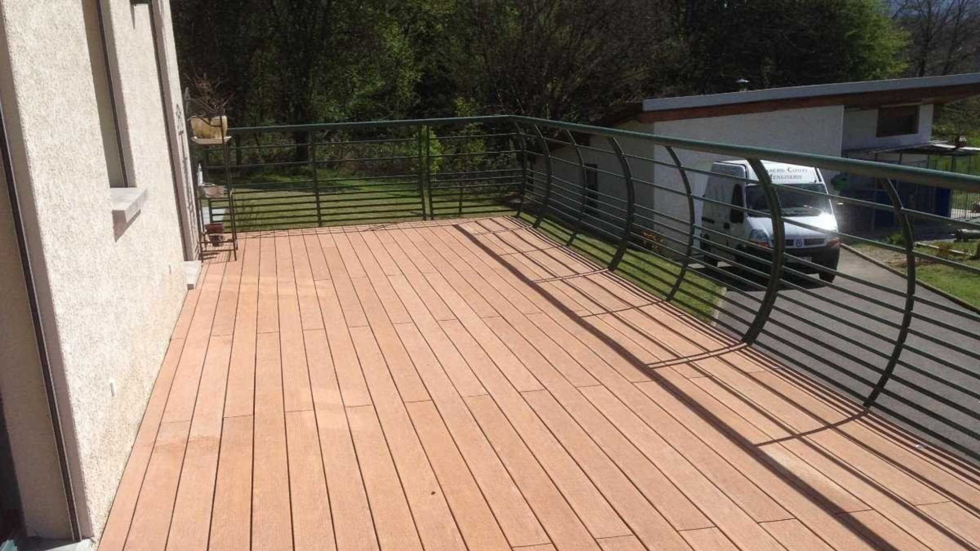 Terrasse en bois avec balustrade noire incurvée, à côté d'un bâtiment et d'arbres. Une camionnette est garée à proximité.