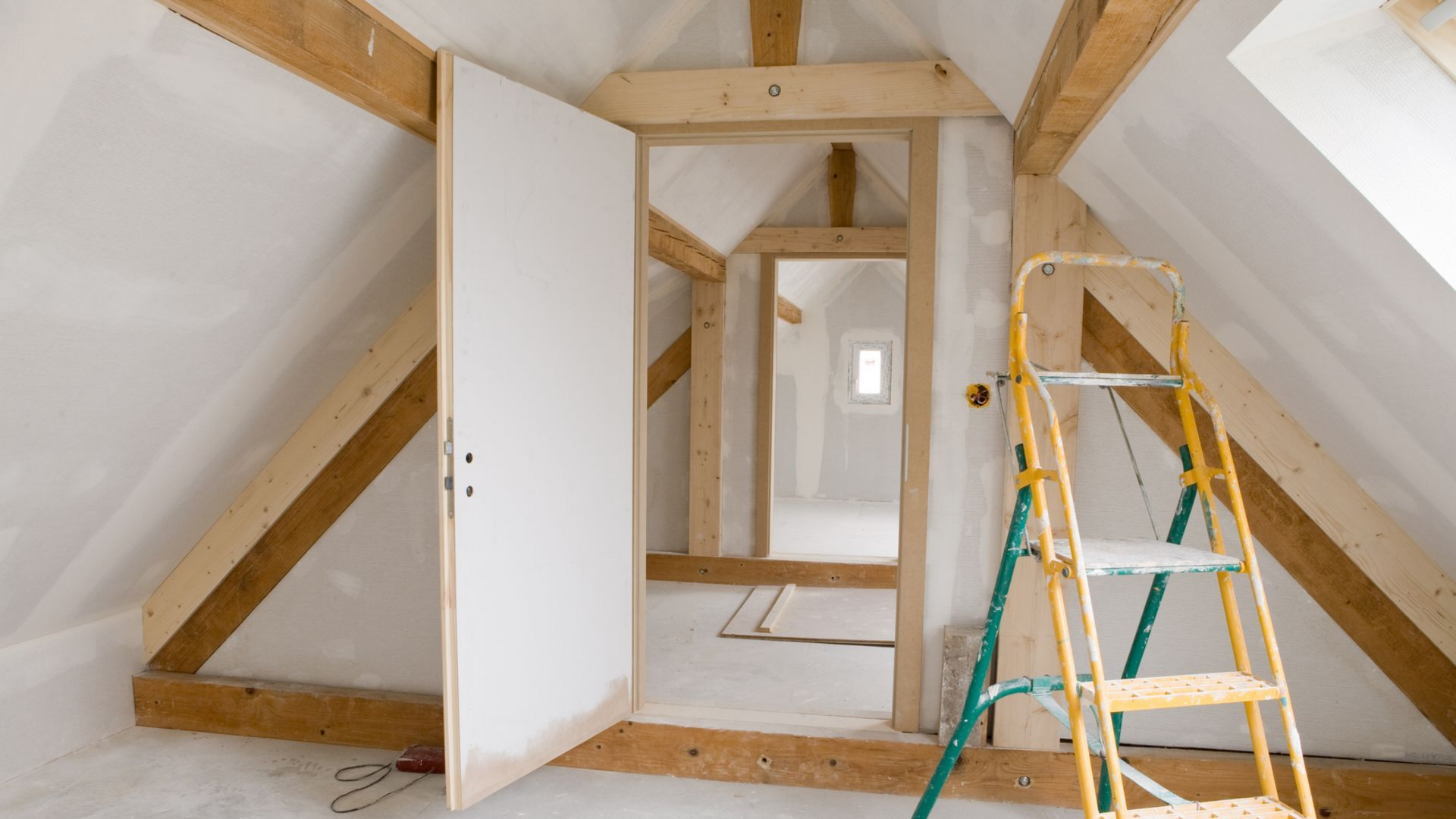 Rénovation des combles en cours ; porte et échelle ouvertes, des poutres en bois, murs et porte blancs.