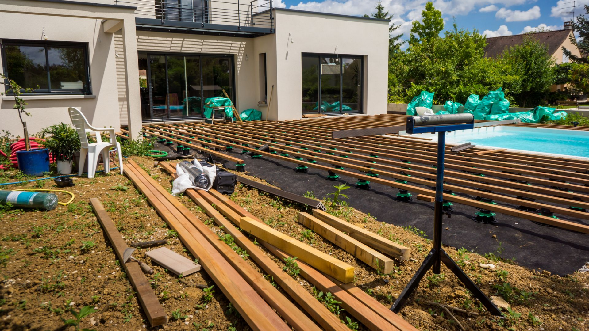 Construction d'une terrasse en bois à côté d'une maison et d'une piscine ; outils et matériaux sont éparpillés sur le sol.