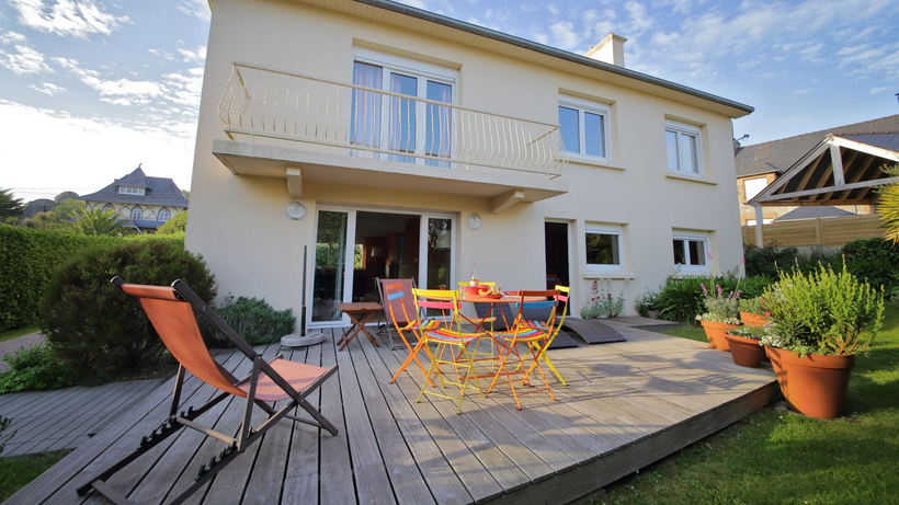 Maison beige à deux étages avec terrasse en bois, mobilier de jardin et chaise de jardin orange sous un ciel bleu.
