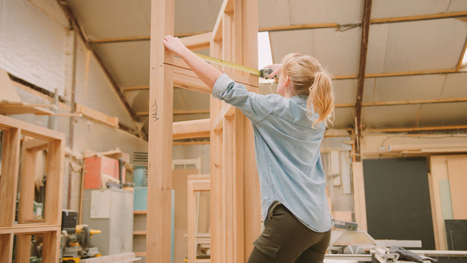 Une femme dans un atelier mesure un cadre en bois avec un ruban à mesurer.