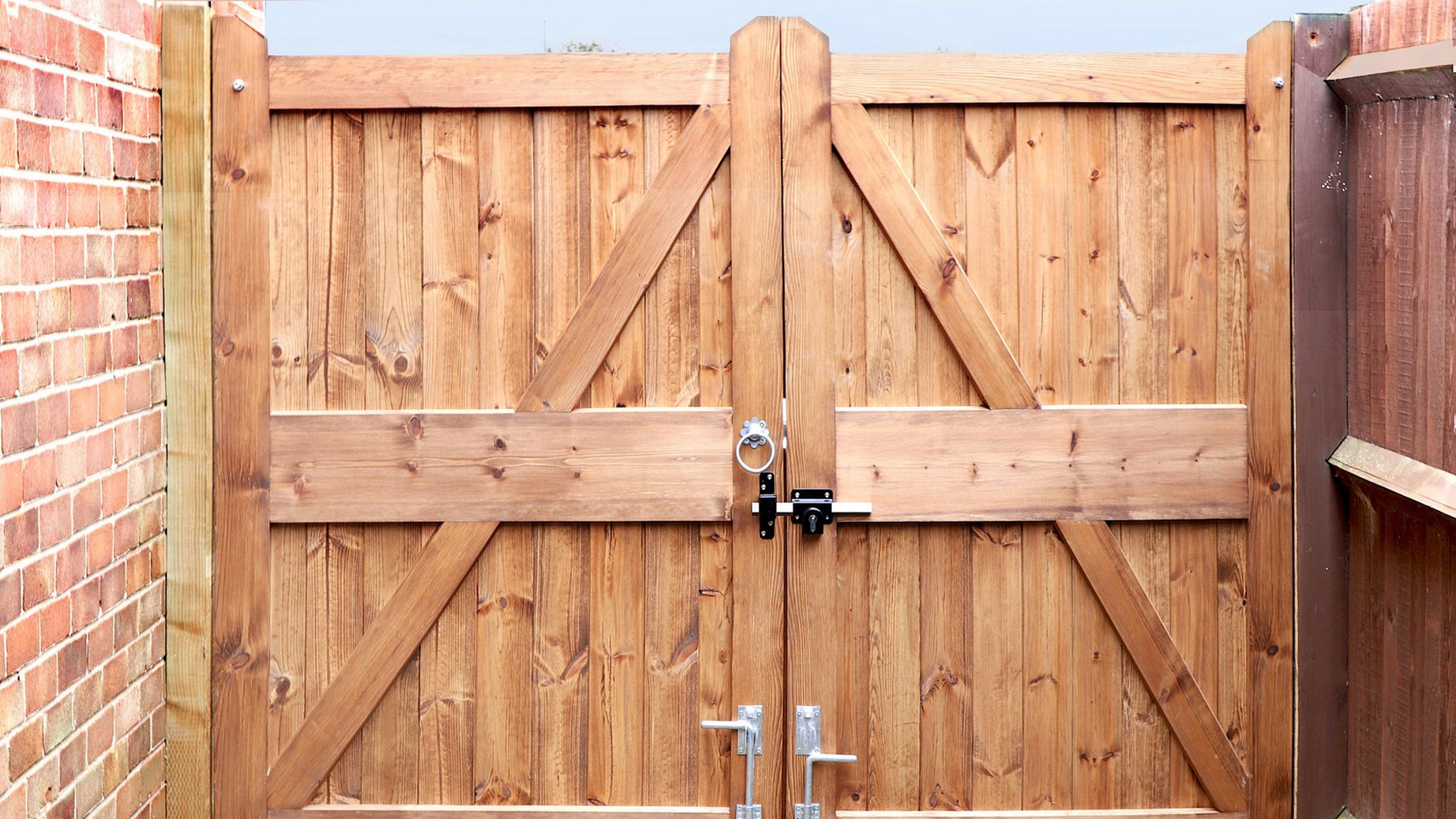 Portail double en bois avec supports diagonaux et loquet, entre un mur de briques et une clôture.