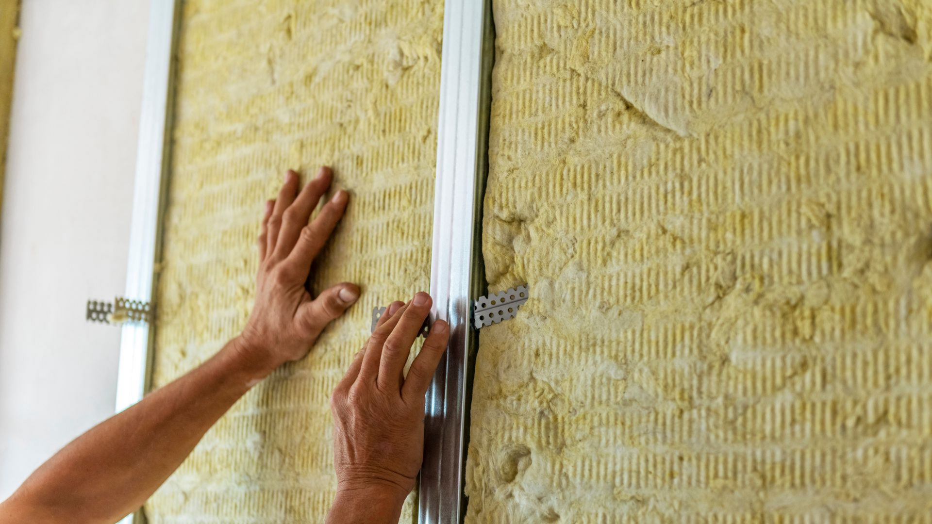 Mains installant une ossature métallique sur un mur rempli d'un isolant jaune.