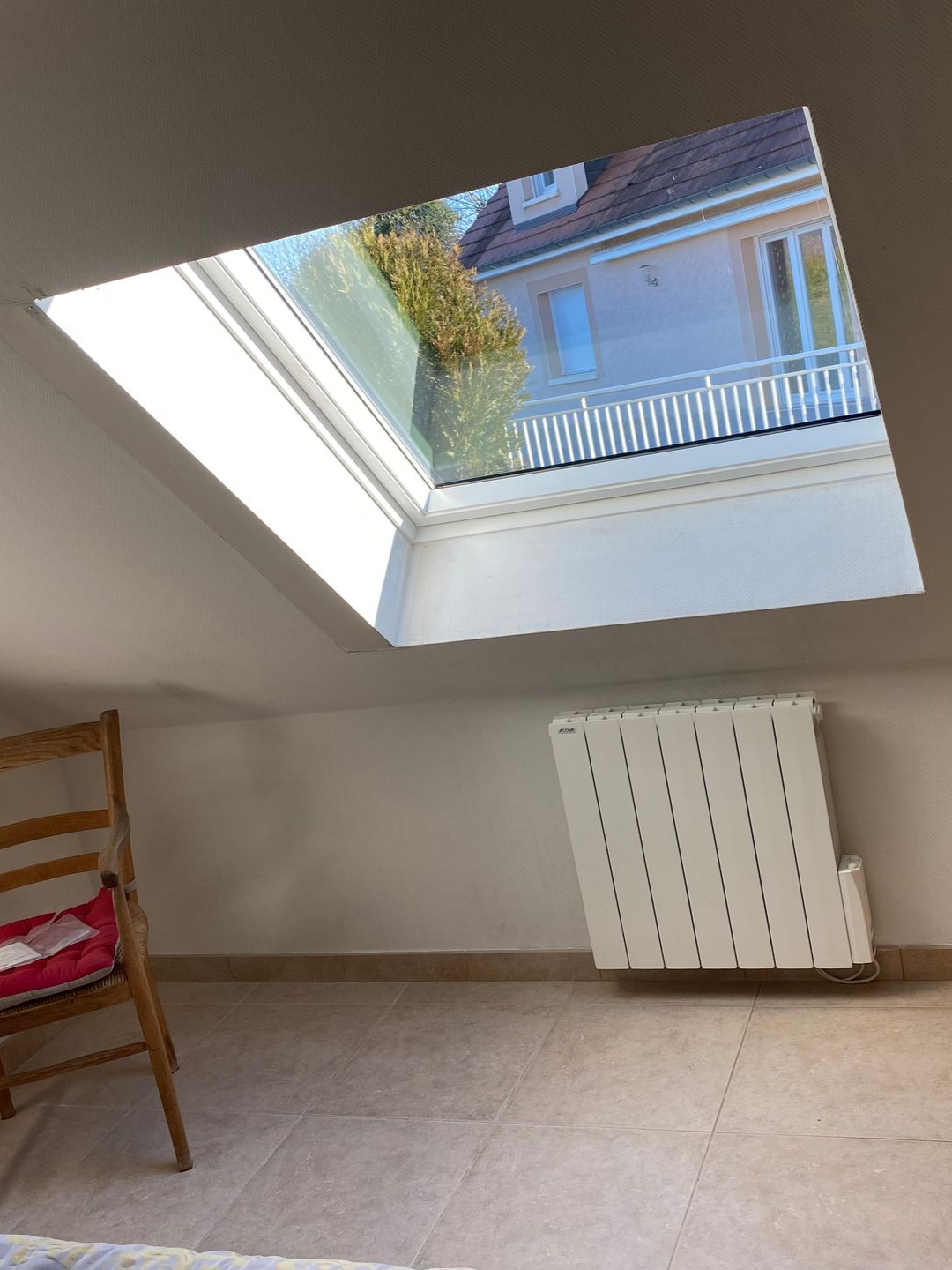 Une fenêtre de toit dans une pièce, avec un radiateur et une chaise en dessous, vue sur un bâtiment à l'extérieur.