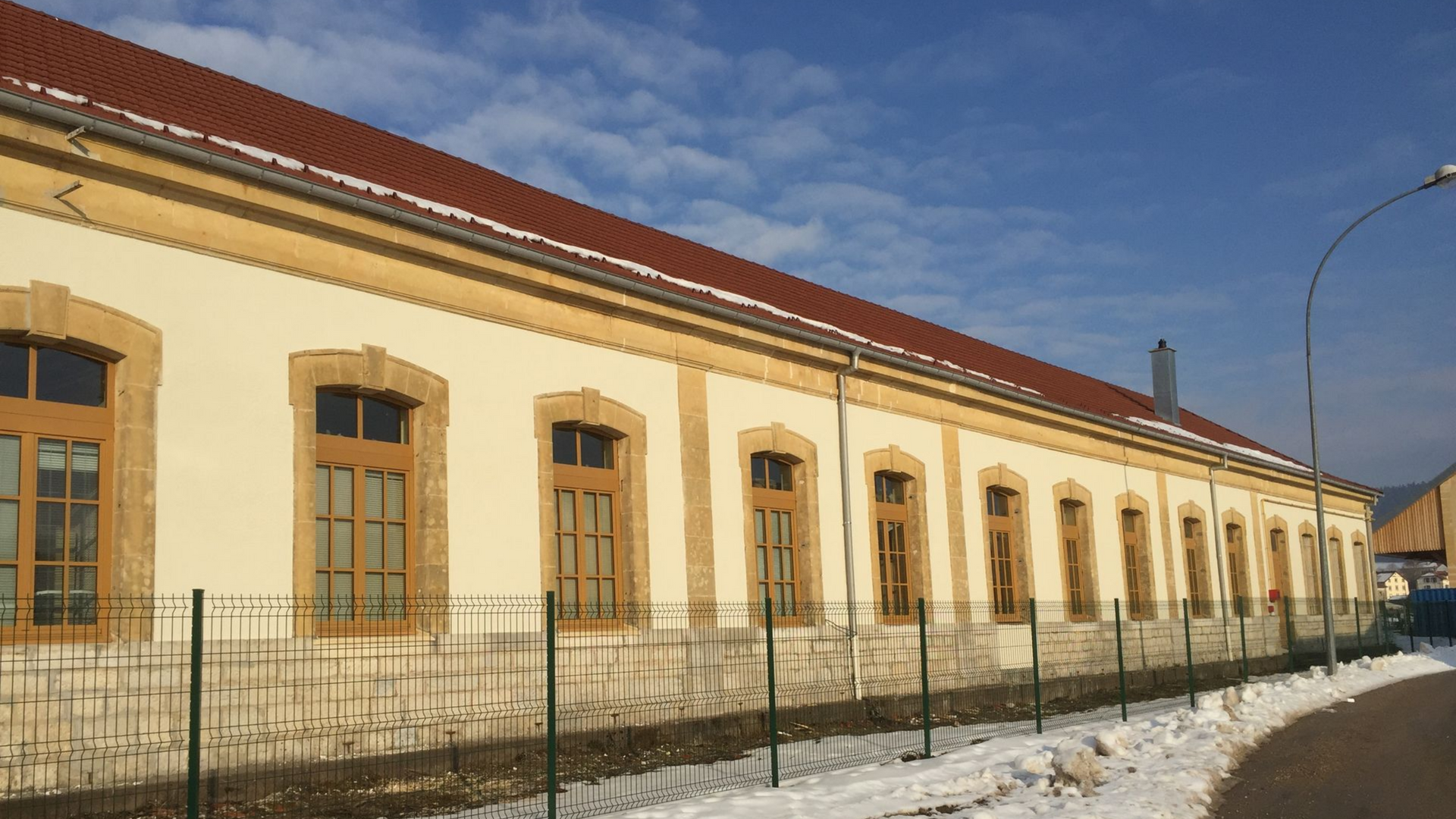 Long bâtiment beige aux fenêtres brunes et au toit rouge, sol enneigé.