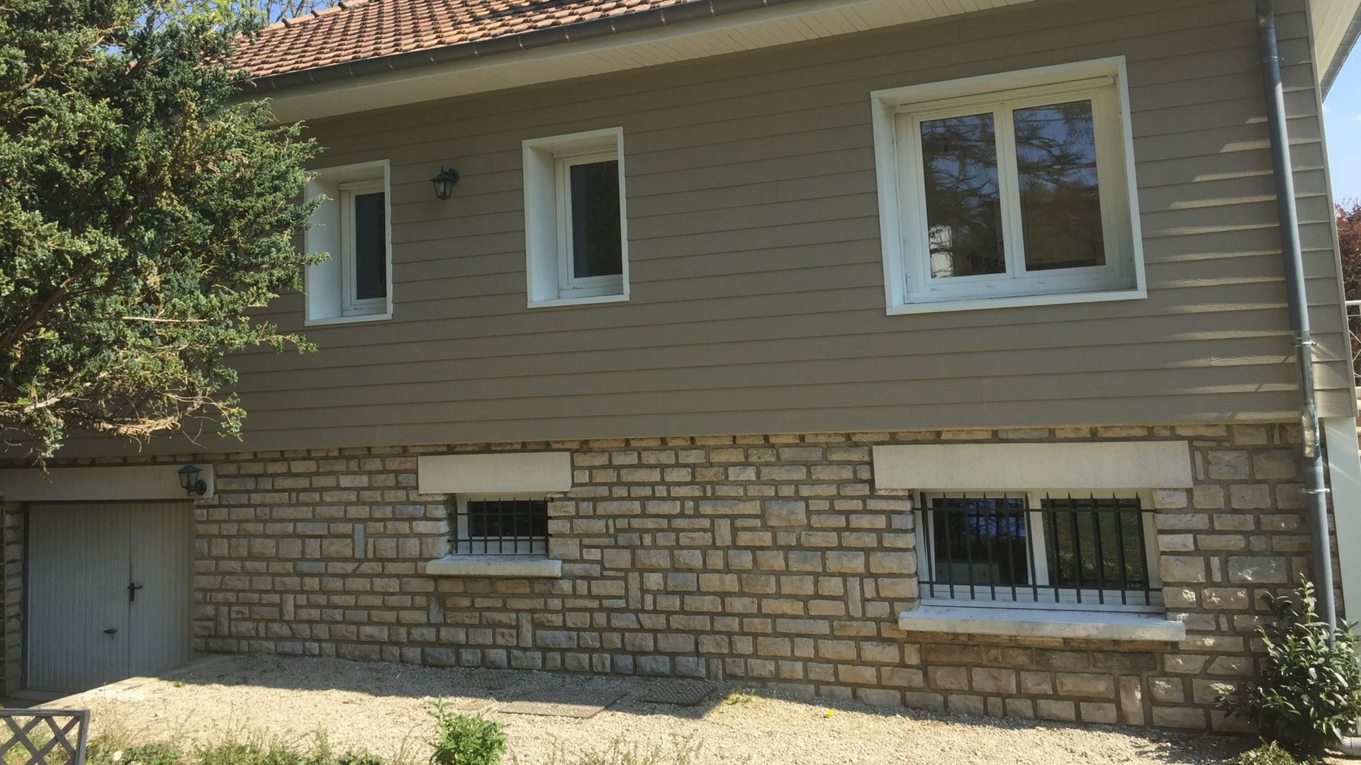 Maison à deux étages avec façade en pierre en bas et revêtement beige en haut ; fenêtres à cadre blanc.