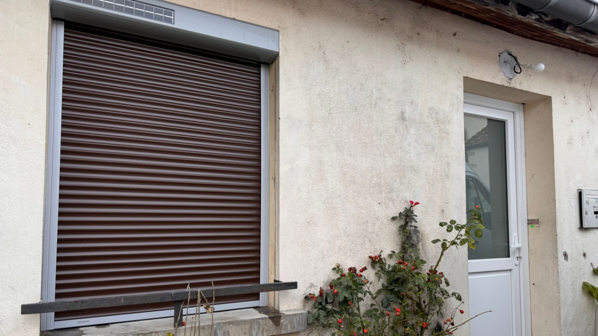 Volet roulant marron fermé sur une fenêtre à côté d'une porte blanche avec un buisson poussant en dessous.