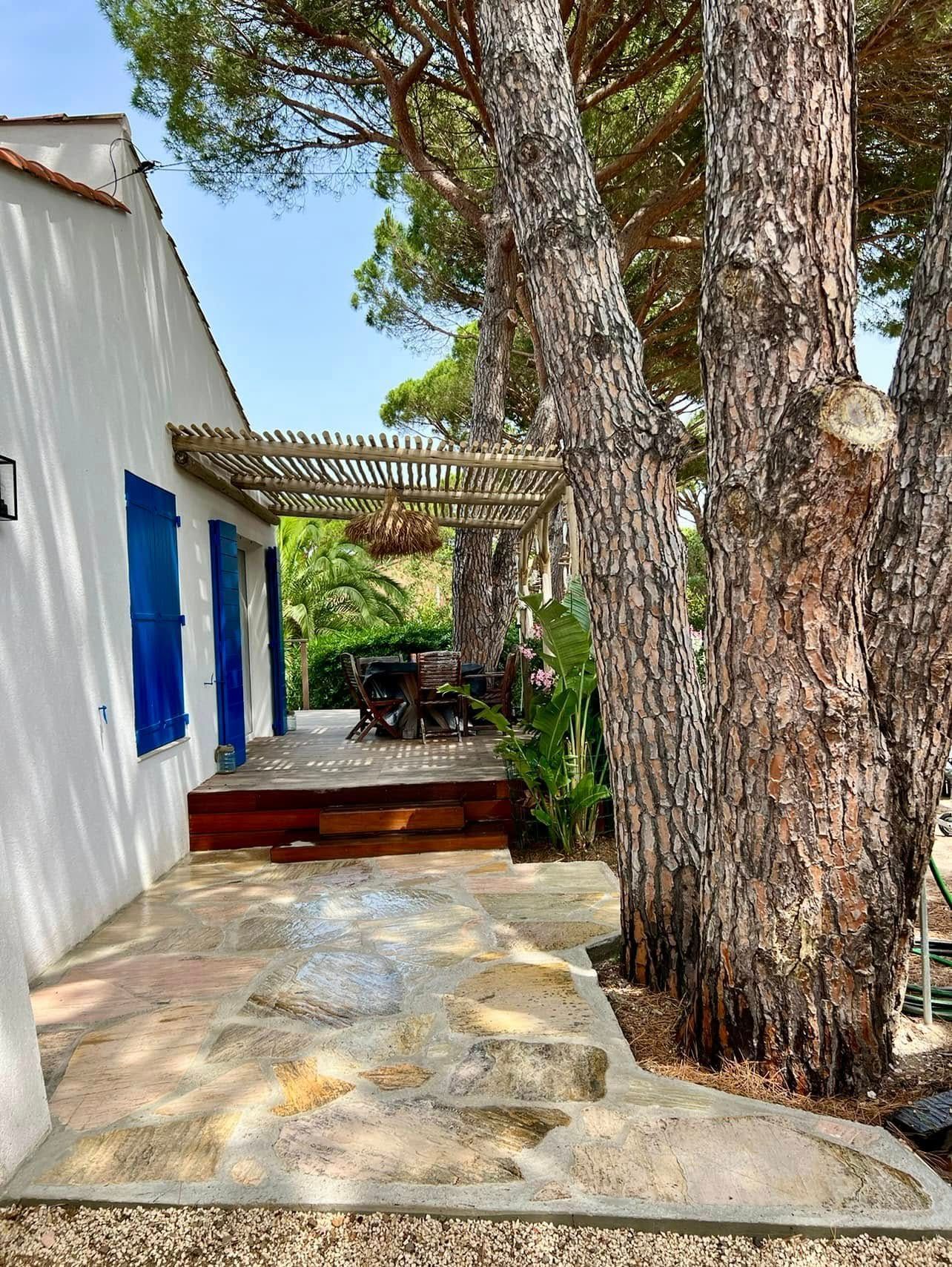 Une terrasse en bois à l'ombre d'une pergola devant une maison aux volets bleus