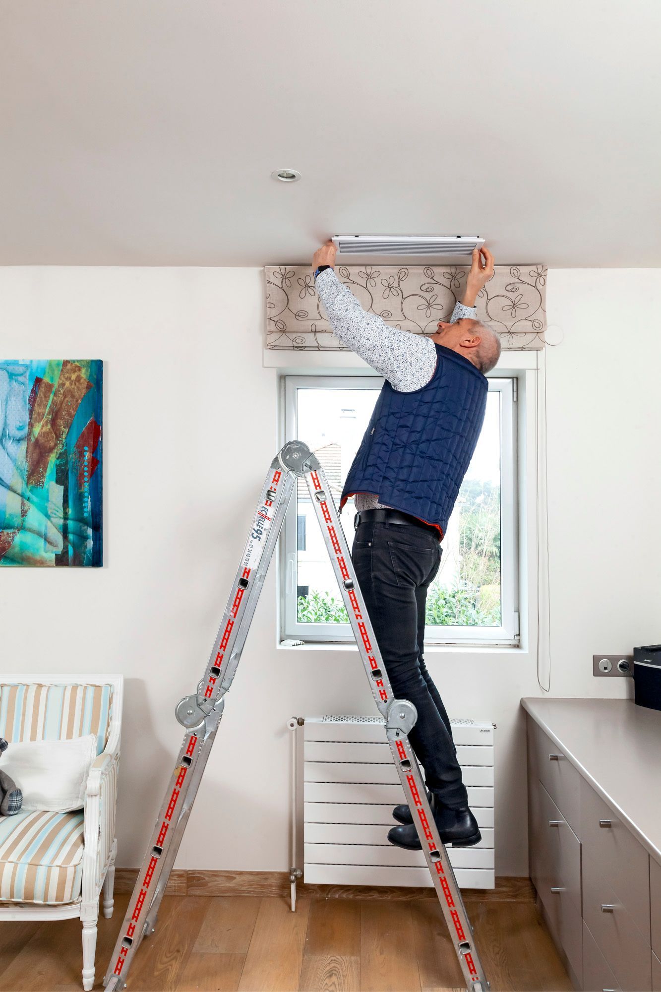 Un homme sur une échelle installe une grille d'aération dans une pièce avec une fenêtre.