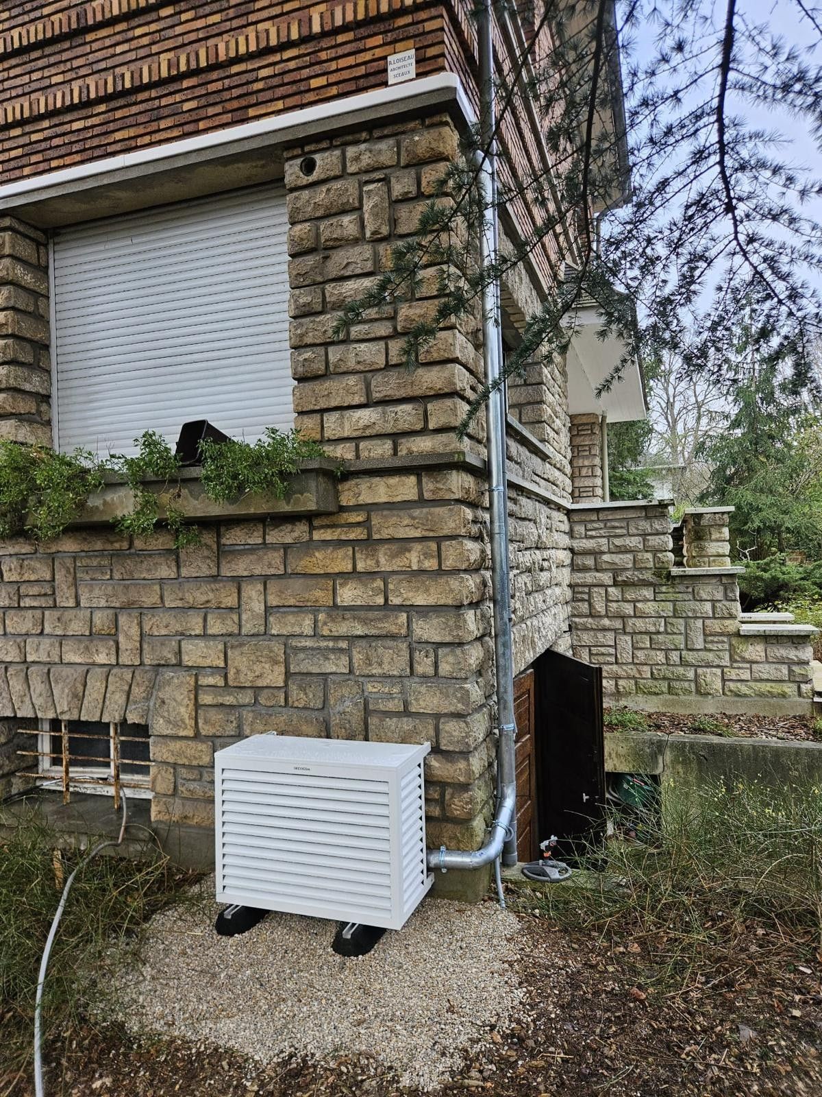 Un climatiseur blanc est installé devant une maison en briques.