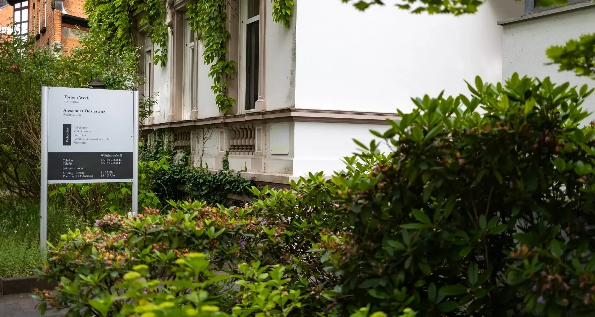 Ein weißes Gebäude einer Kanzlei mit einem Schild davor, umgeben von Bäumen und Büschen.