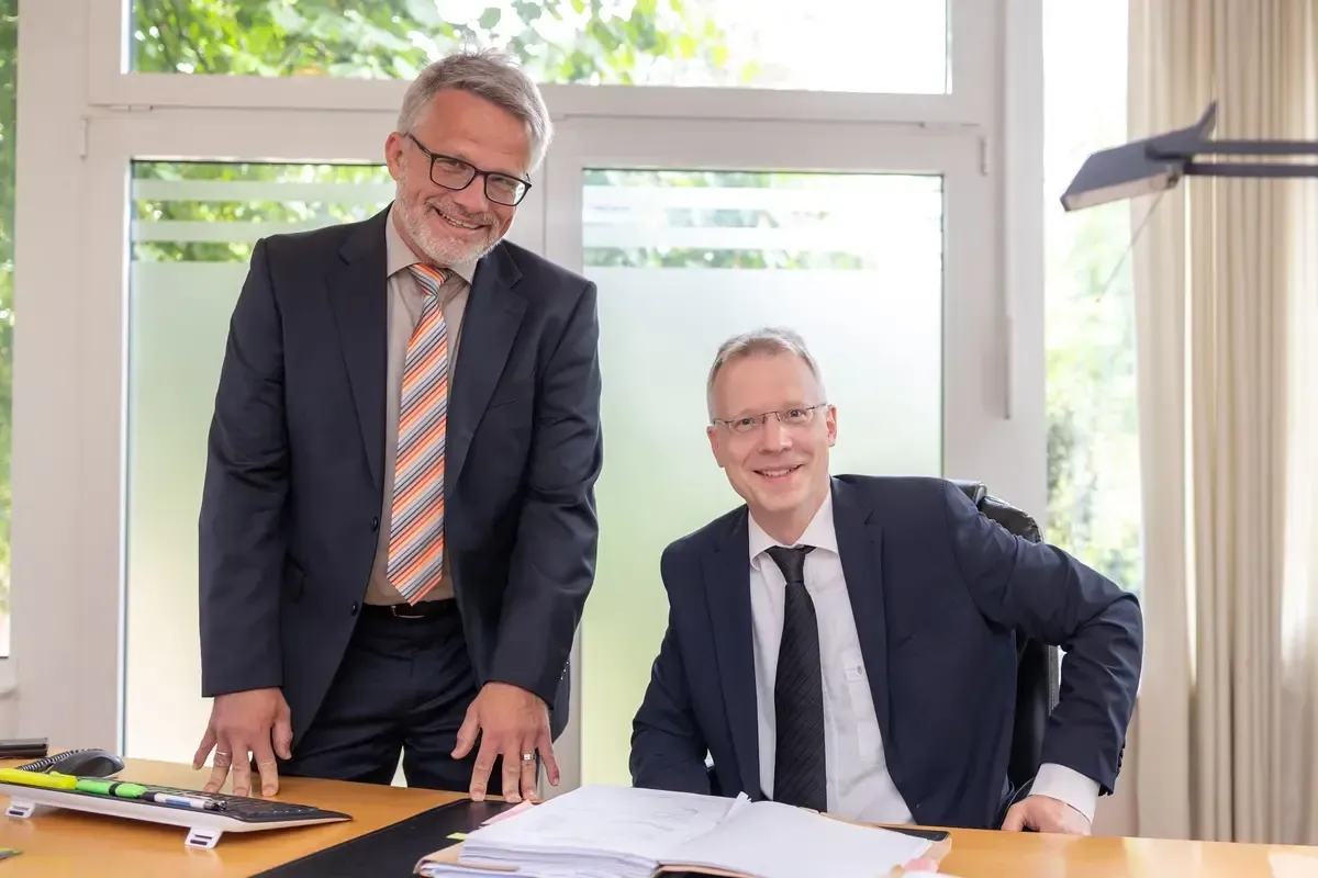 Zwei Anwälte in Anzug und Krawatte stehen nebeneinander vor einem Schreibtisch.