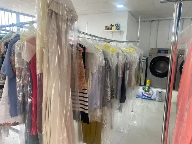 Ropa tendida en una tintorería, cubierta de plástico. Se ve una lavadora al fondo.