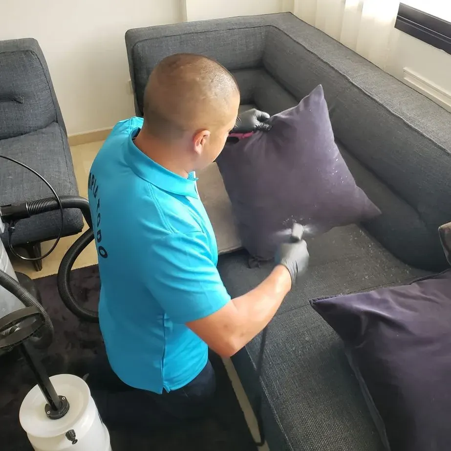 Un hombre con una camisa azul está limpiando un sofá con una aspiradora.
