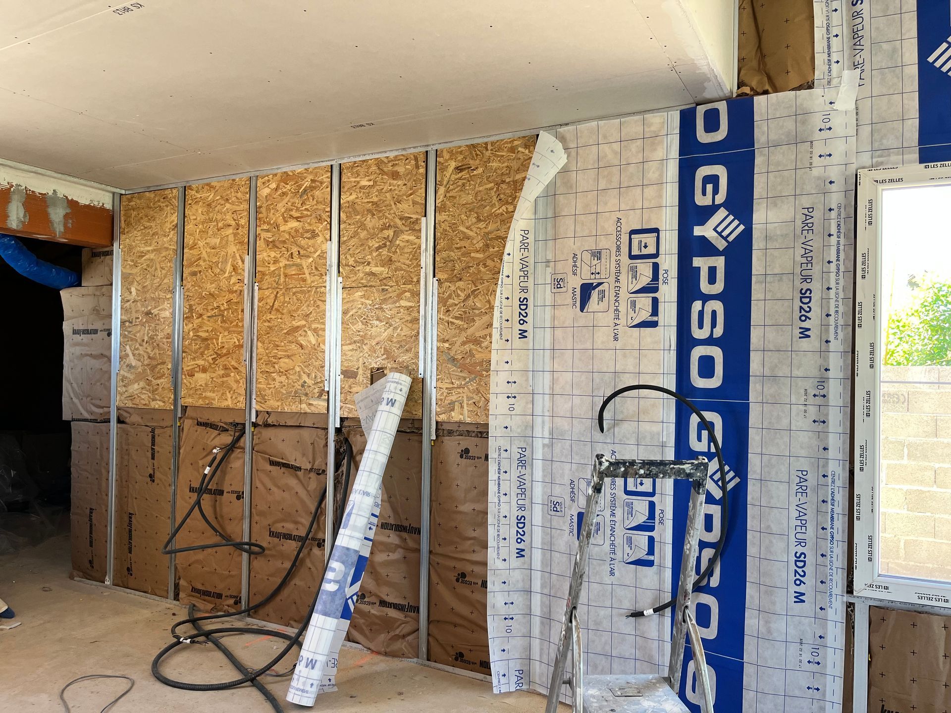 Pièce partiellement aménagée avec isolation apparente, ossature bois et panneaux de plaques de plâtre contre le mur.