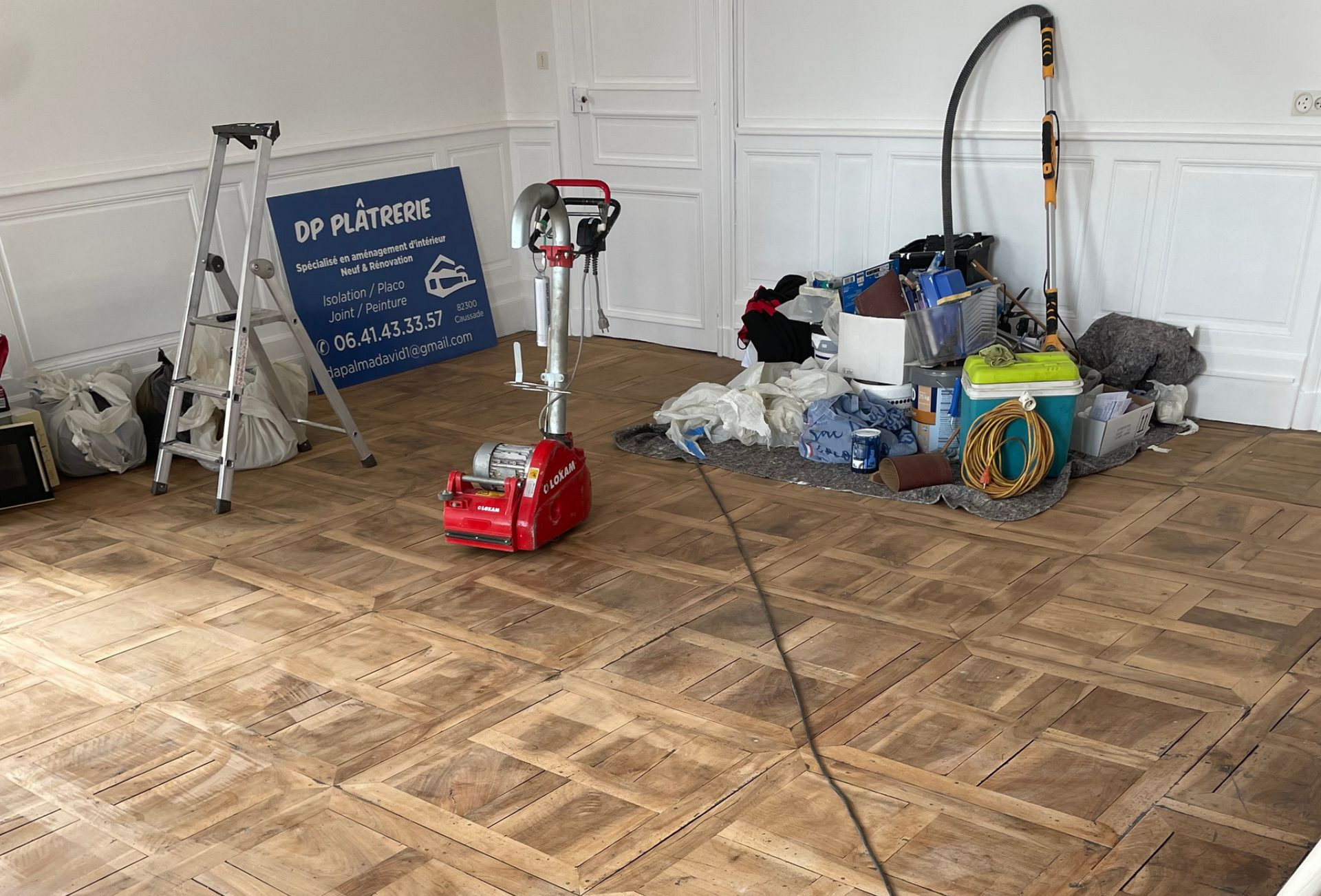 Un parquet est en cours de rénovation dans une pièce vide, avec des outils et des fournitures de ponçage éparpillés un peu partout.