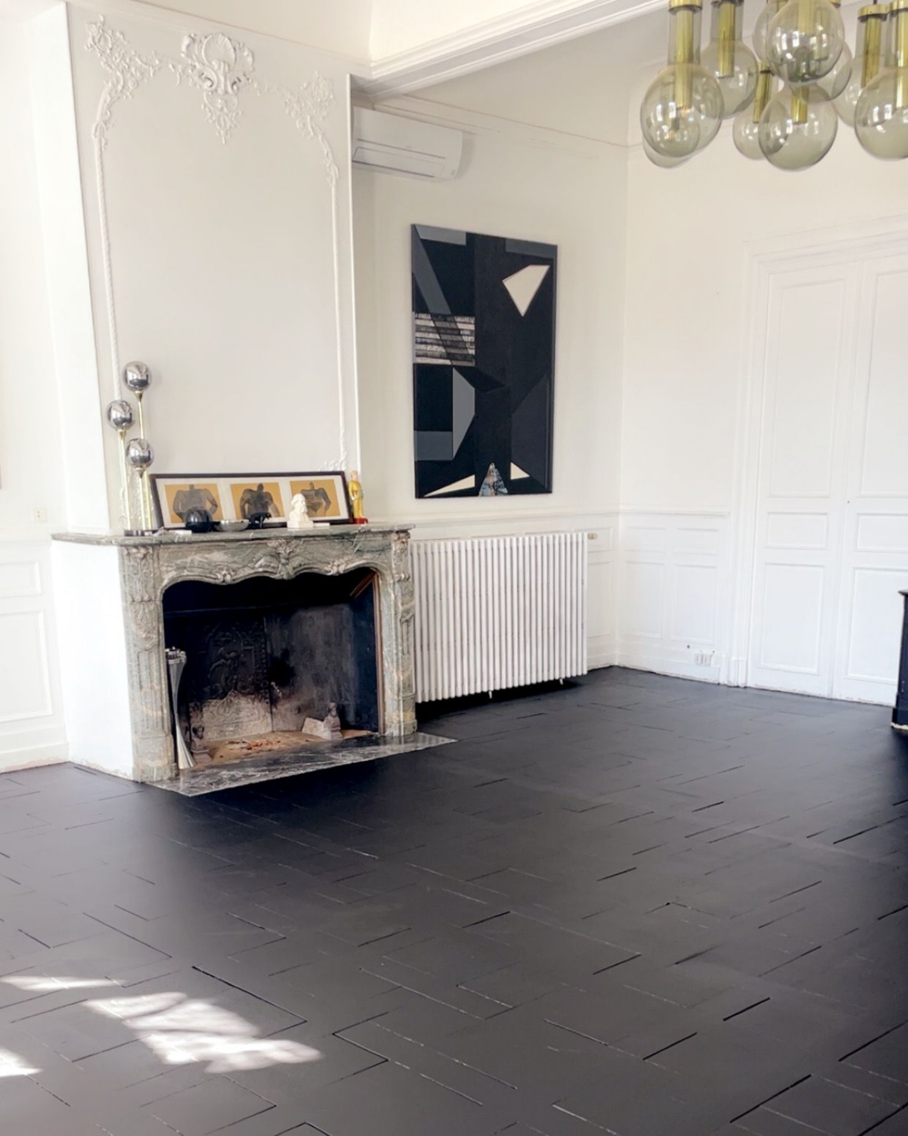 Salon lumineux d'un blanc éclatant avec sol noir, cheminée, radiateur et grande fenêtre laissant entrer la lumière du soleil.