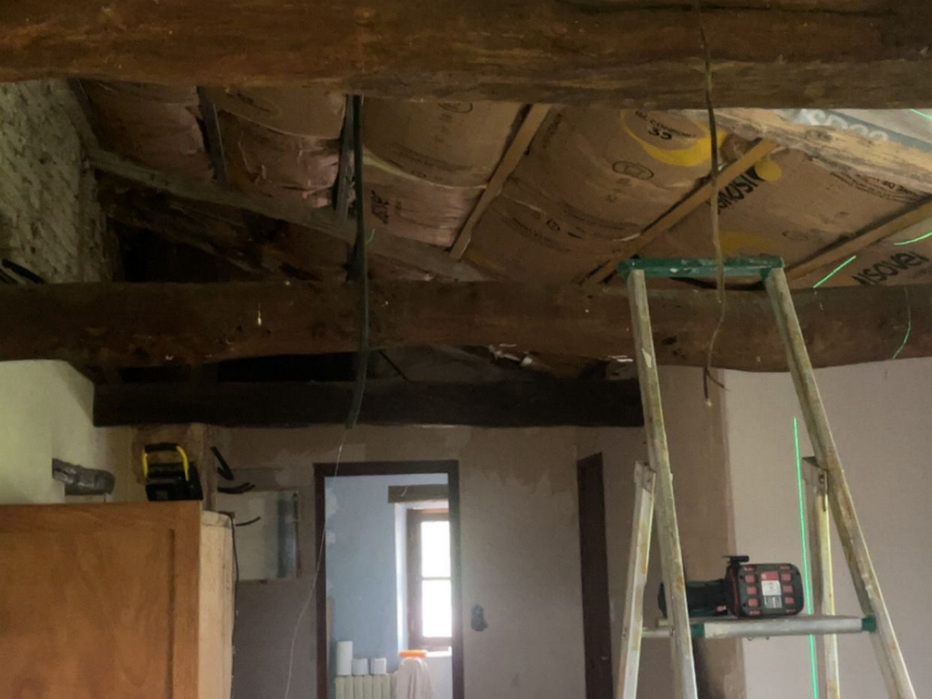 Pièce inachevée avec isolation du plafond apparente, échelle et porte donnant sur un espace plus lumineux.