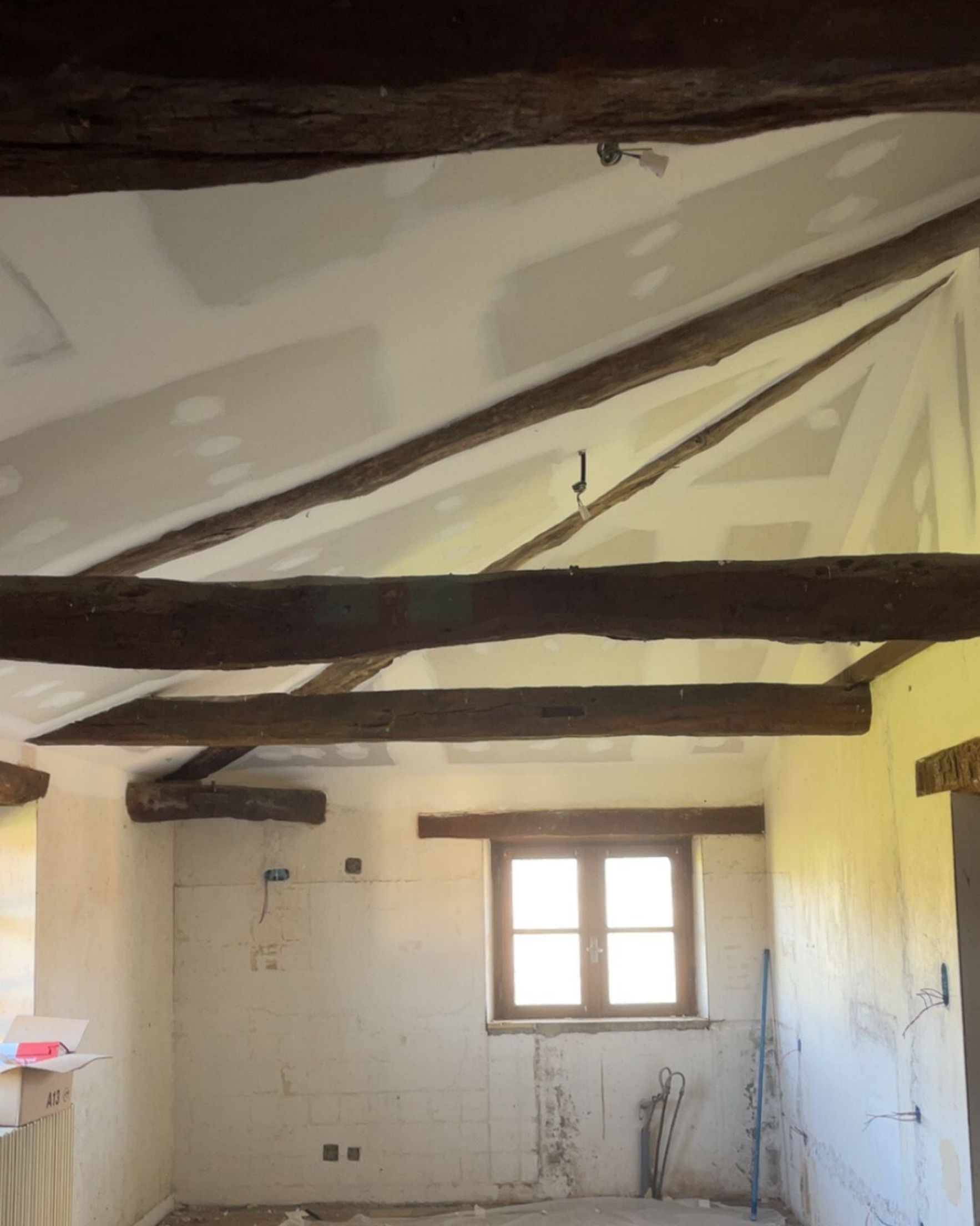 Chambre partiellement rénovée avec poutres apparentes, murs blancs et une petite fenêtre au fond.