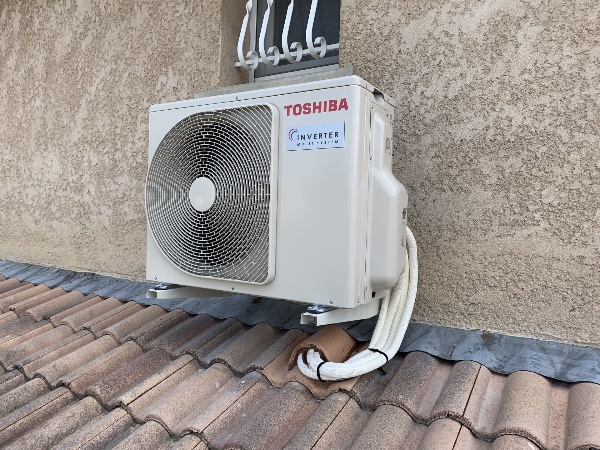 Unité extérieure de climatiseur Toshiba blanche, fixée au mur à côté des tuiles du toit, avec des tuyaux gainés en dessous.