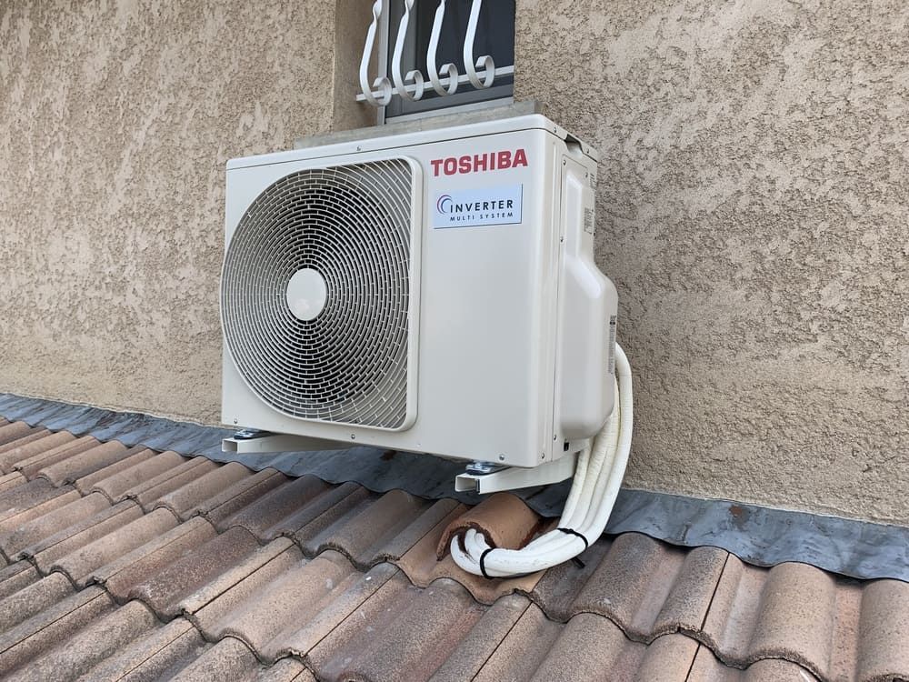 Unité de climatisation Toshiba blanche, montée sur un mur extérieur en stuc au-dessus d'un toit de tuiles.