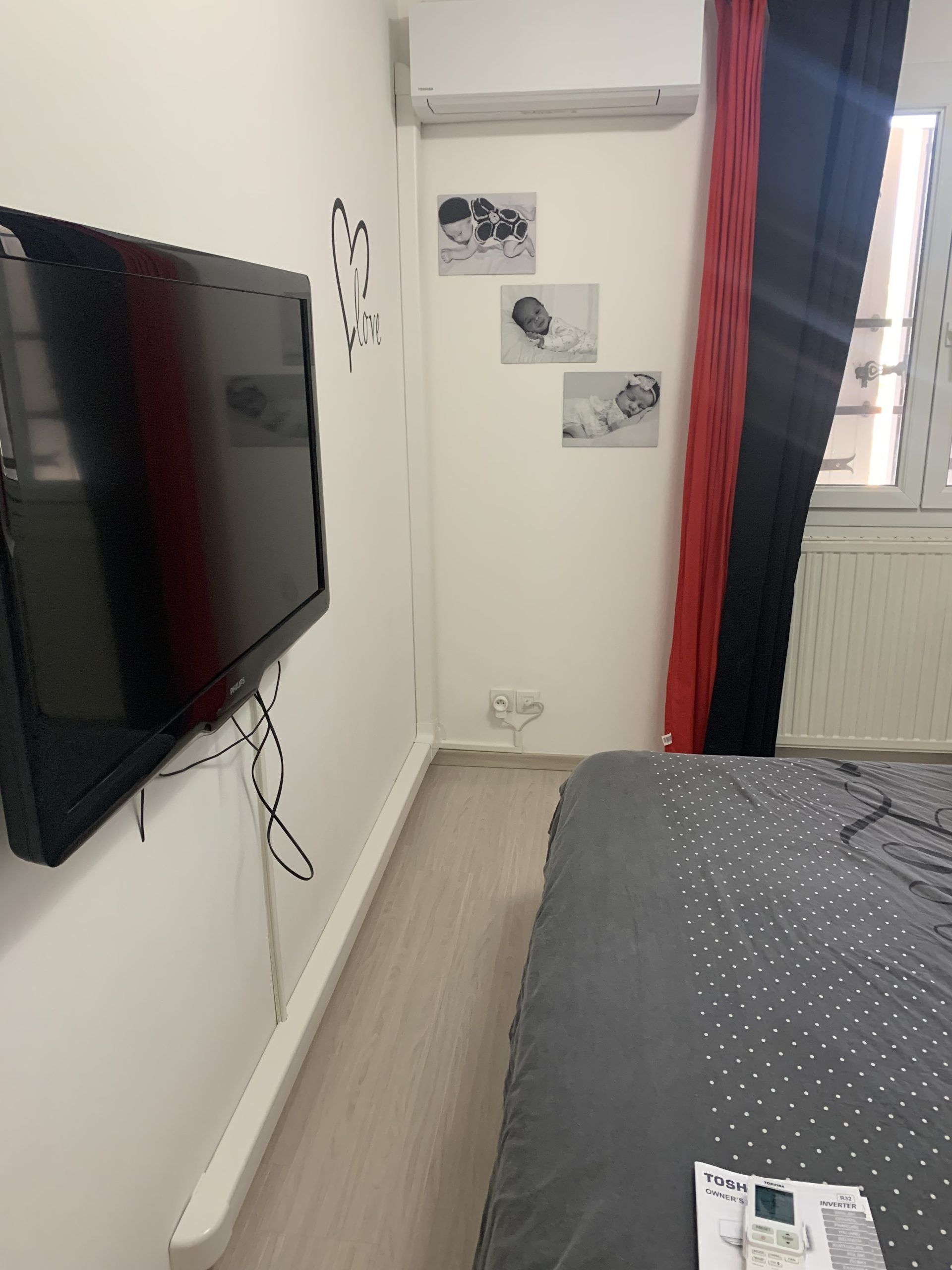 Chambre avec télévision murale, lit et petites photos encadrées sur un mur blanc près d'une fenêtre.