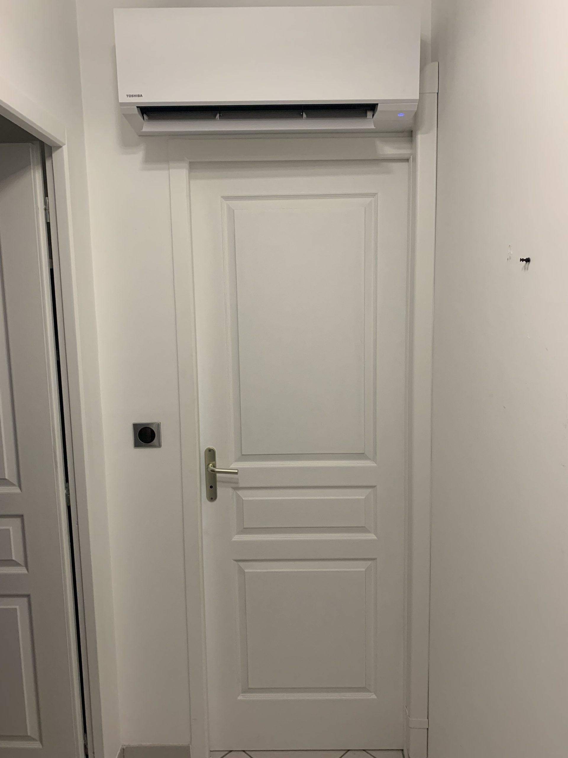 Porte intérieure blanche avec poignée en laiton et climatiseur mural au-dessus, dans un couloir étroit