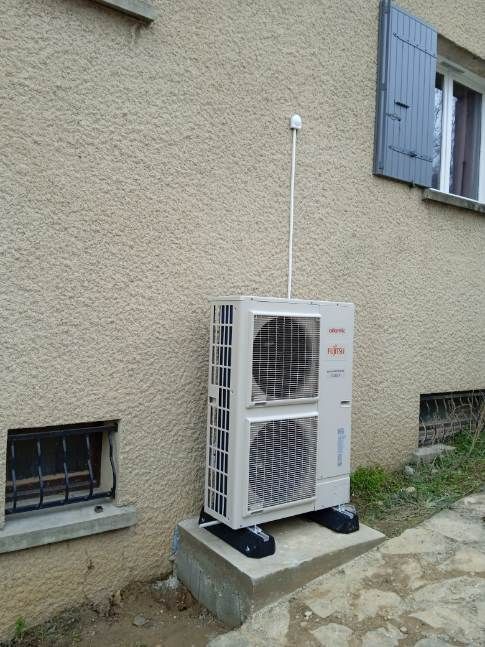 Unité de climatisation extérieure adossée à un mur beige, avec un ventilateur visible et un mât blanc ressemblant à une antenne au-dessus.