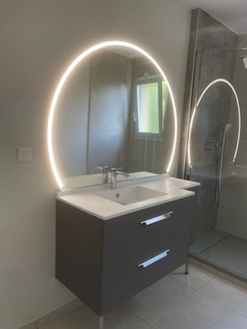 Meuble-lavabo moderne avec miroir ovale rétroéclairé, lavabo blanc et armoires foncées à côté d'une douche en verre.