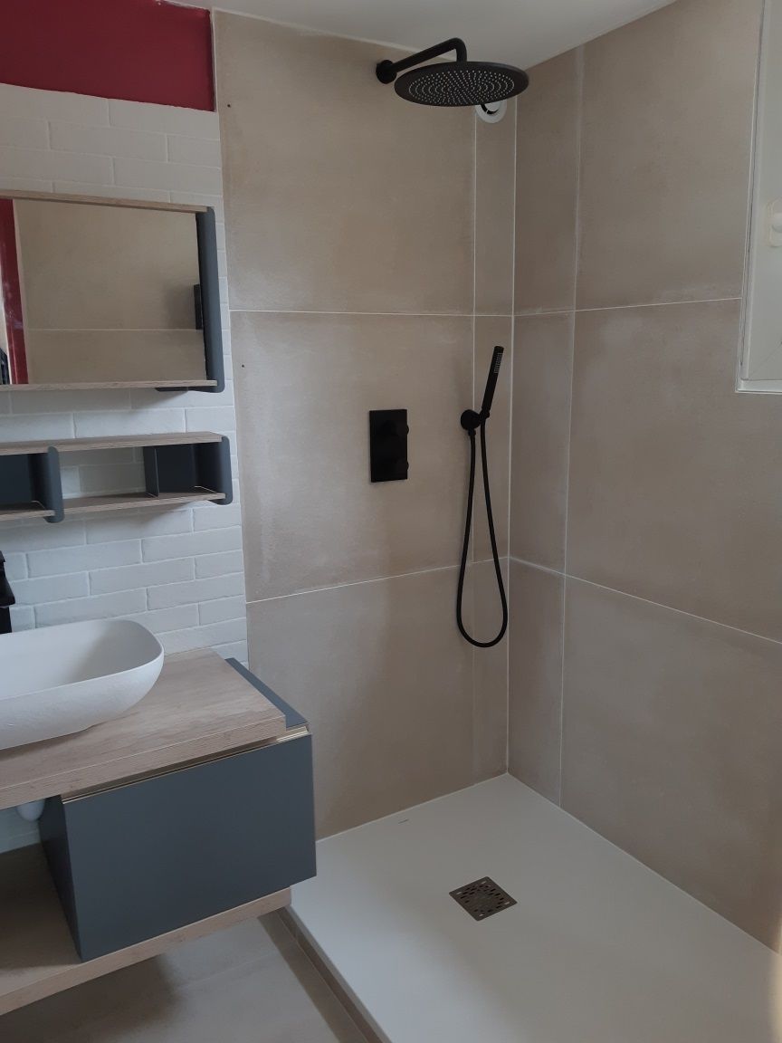 Douche moderne dans une salle de bain avec carrelage beige, robinetterie noire et meuble vasque sur la gauche.
