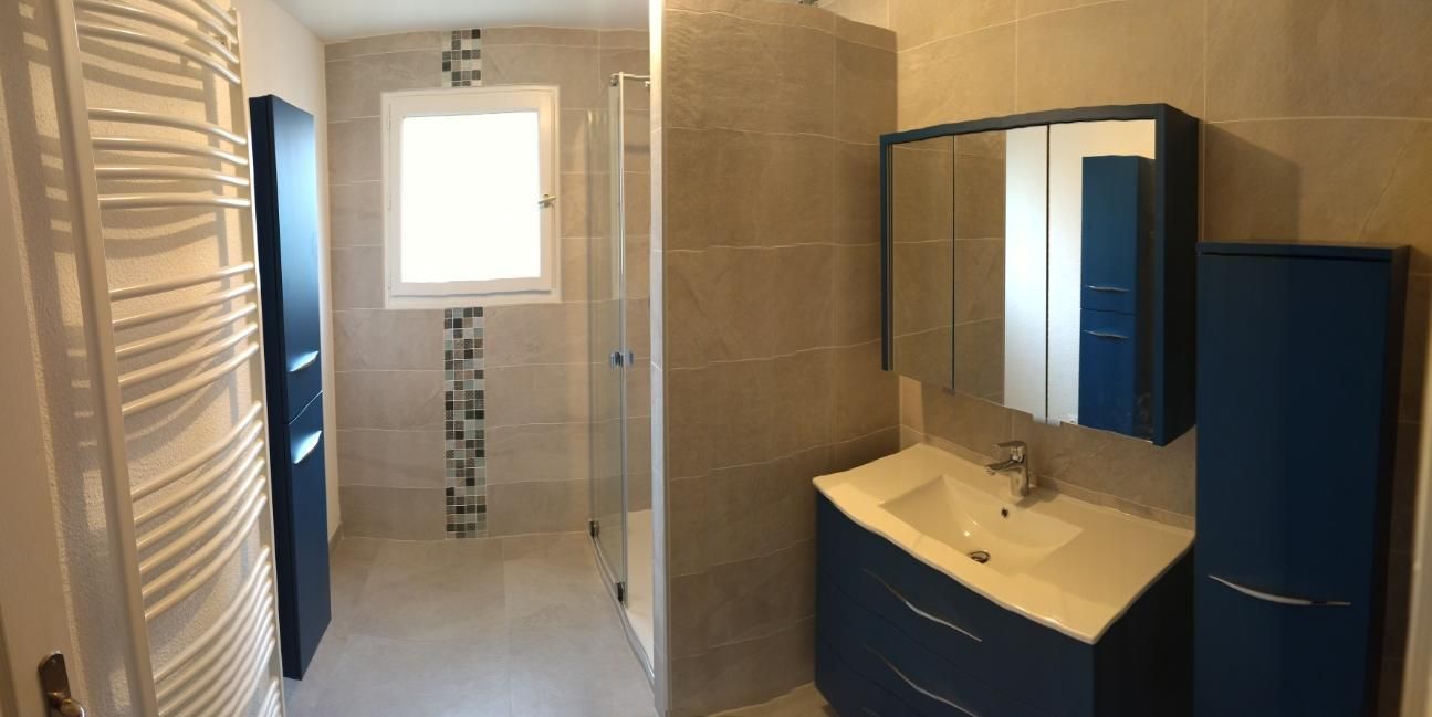 Salle de bains étroite avec carrelage beige, douche à gauche, fenêtre en face et meuble-lavabo bleu avec miroir à droite.