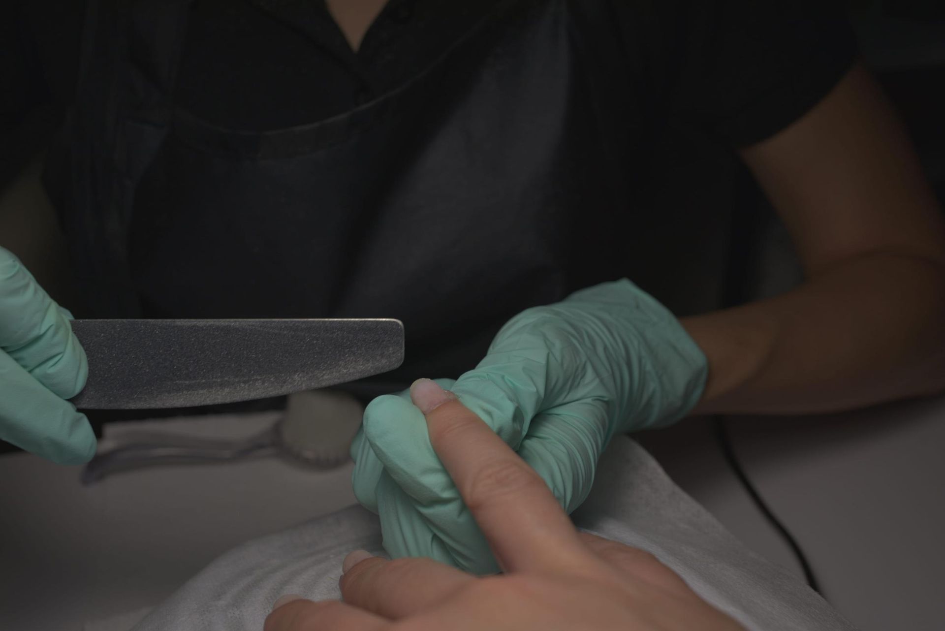 Manicura de uñas con lima y herramienta para cutículas sobre una toalla blanca; se ven los guantes color turquesa.