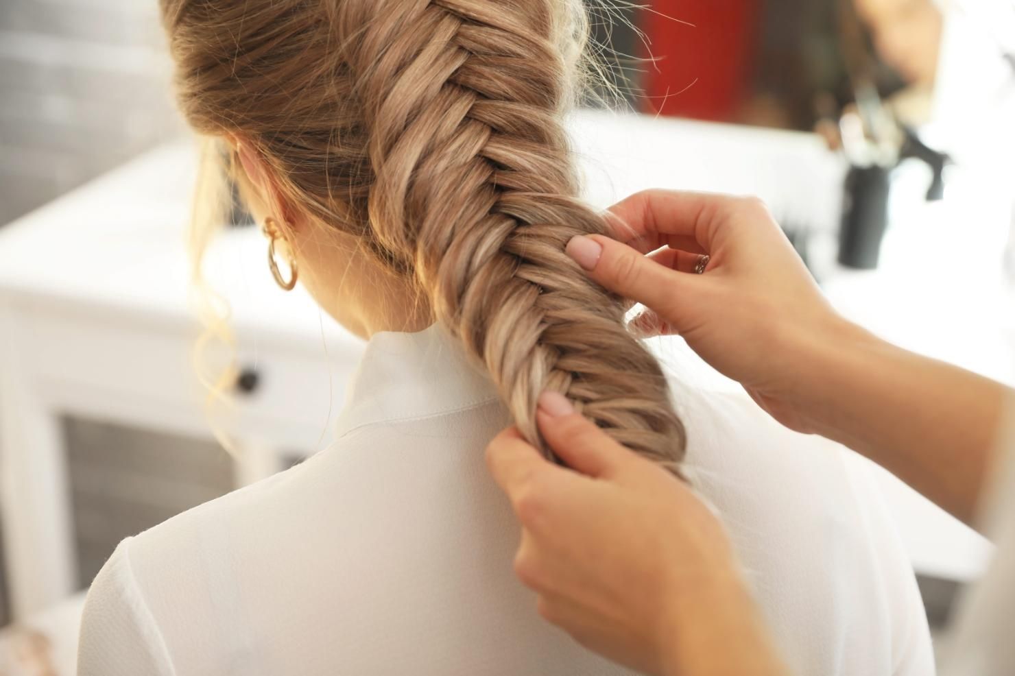 Unas manos trenzan cabello rubio frente a un espejo en una habitación luminosa.