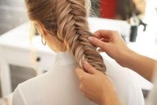 Unas manos trenzan cabello rubio frente a un espejo en una habitación luminosa.