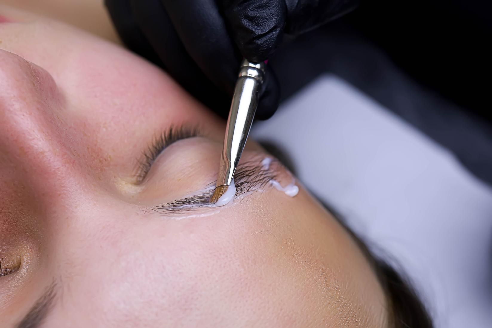 Primer plano de una brocha de maquillaje aplicando producto alrededor de un ojo cerrado durante un tratamiento de pestañas.