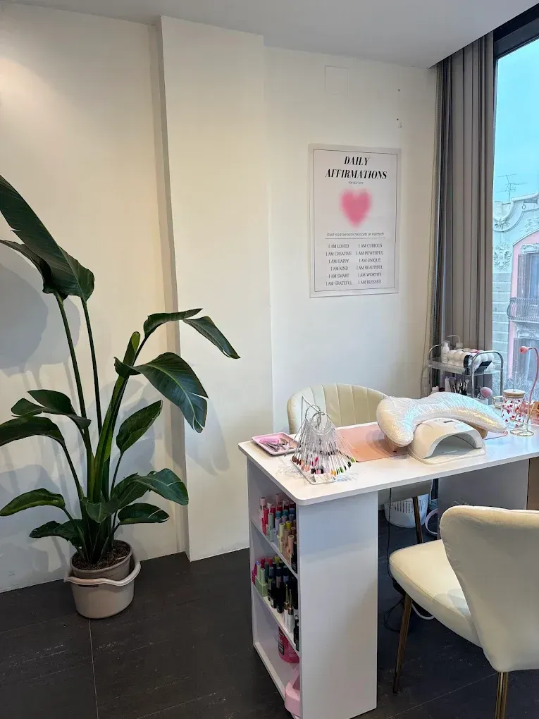 Acogedor rincón de manicura con escritorio blanco, silla color crema, planta en maceta y cuadro rosa junto a la ventana.