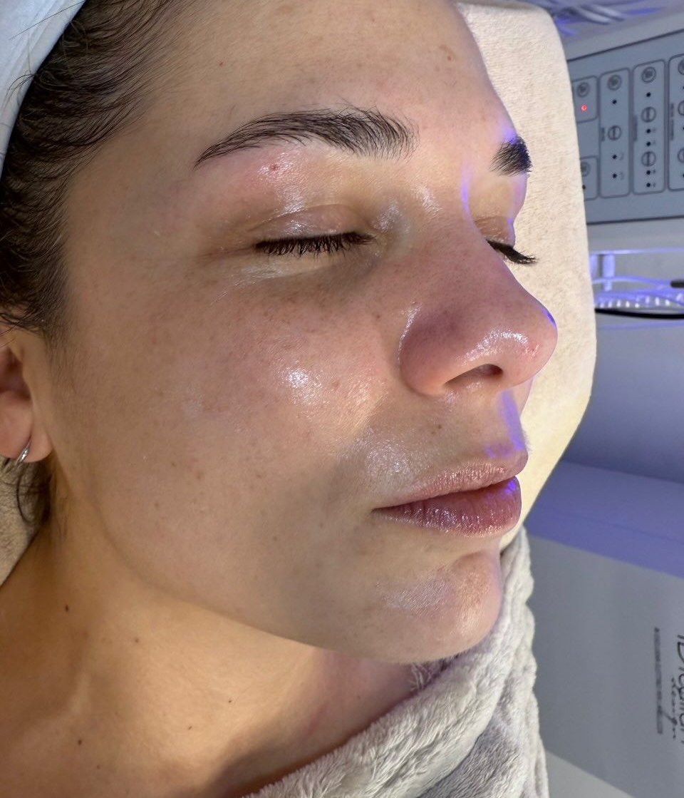Primer plano de una persona con un tratamiento facial brillante, con los ojos cerrados, tumbada en una camilla de spa.