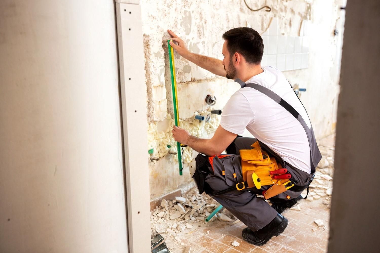 Obrero mide la pared con una cinta métrica; hay escombros en el suelo.