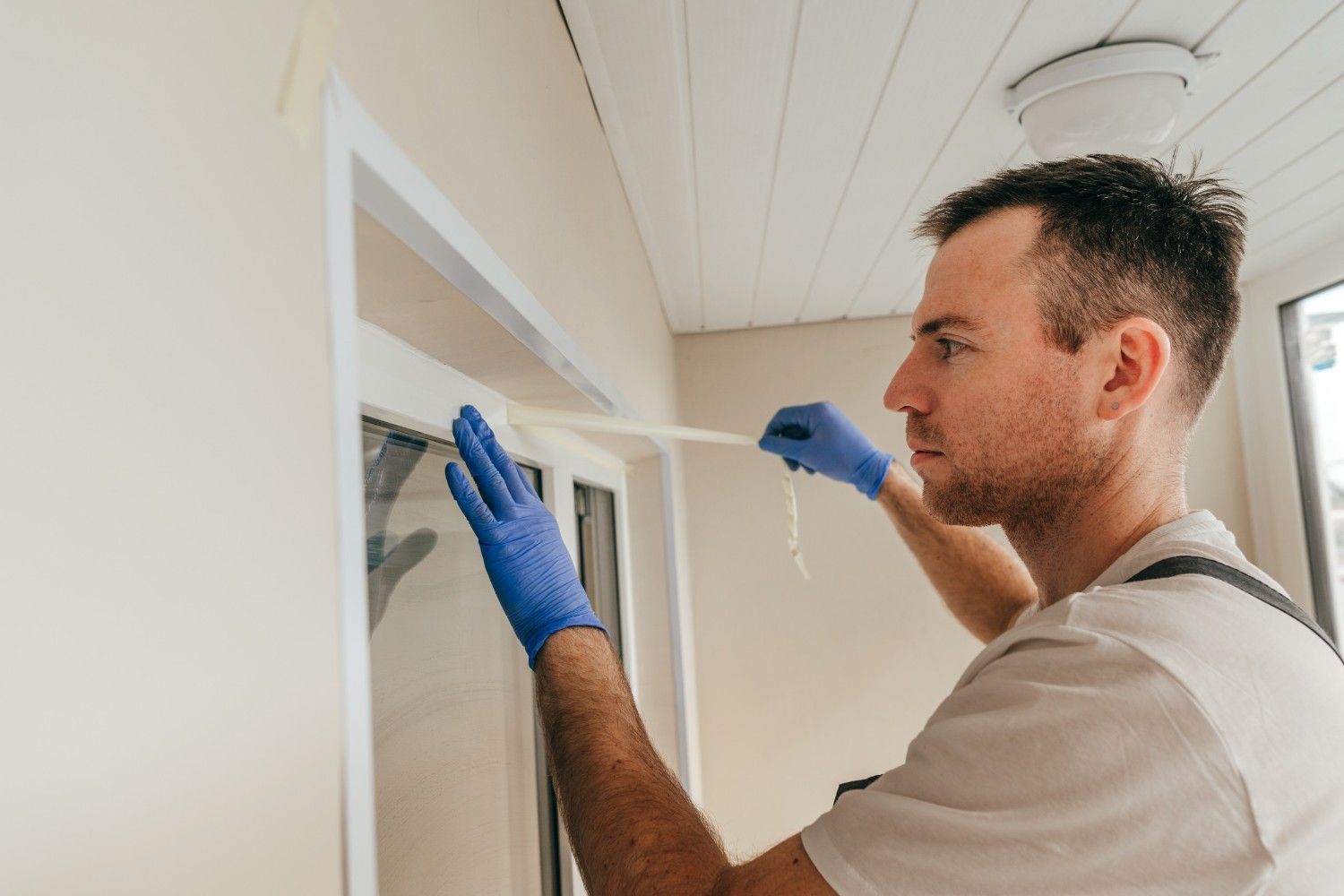 Un hombre con guantes azules aplica cinta adhesiva al marco de una ventana en una pared beige.