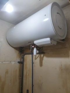 Chauffe-eau blanc fixé au mur dans une pièce avec des canalisations.