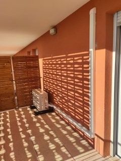 Balcon aux murs orange, agrémenté de panneaux de bois projetant des ombres à motifs. Un climatiseur est posé à même le sol.